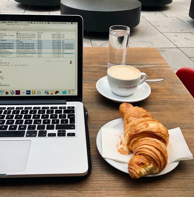 Coworking in Kigali_Living In Kigali_ A Macbook Pro, cappuccino, a water, and a croissant are in the photo. They are sitting outside.